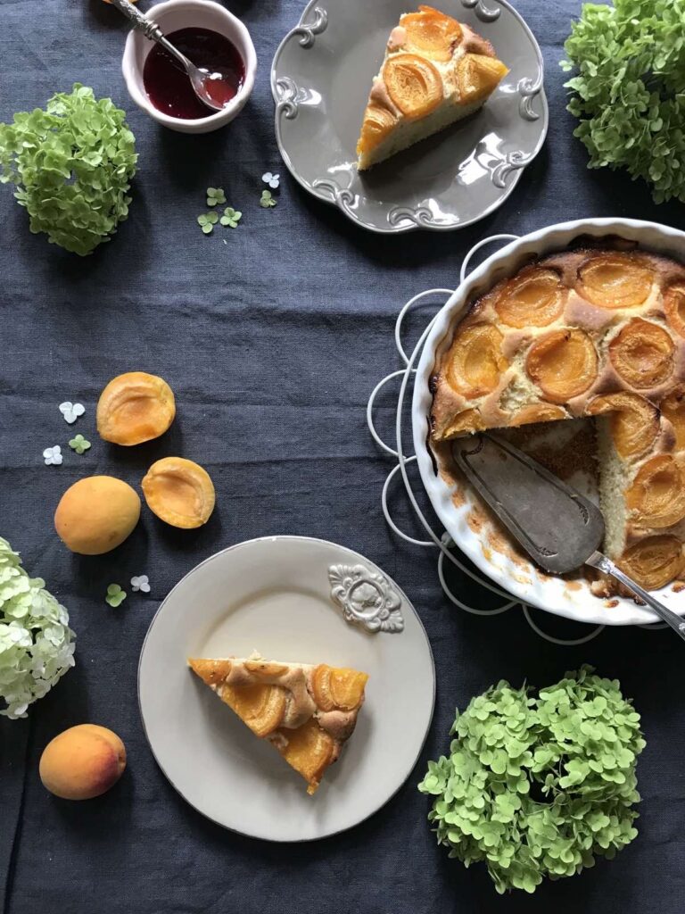 Overhead view of a partially sliced homemade apricot cake in a white baking dish, with two slices served on rustic plates, scattered fresh apricots, a small bowl of jam, and green hydrangeas on a dark blue linen cloth.