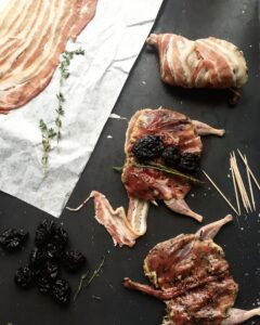 Top-down view of raw quails being stuffed with dried prunes and wrapped in bacon slices, surrounded by toothpicks and fresh thyme on a dark surface.