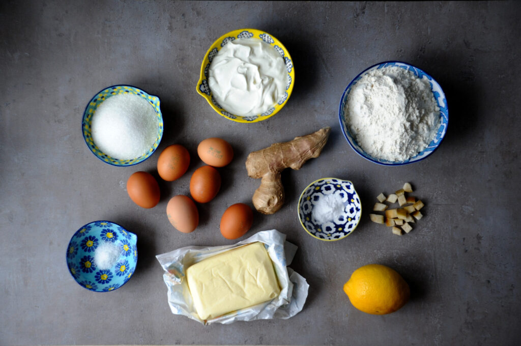 Flat lay of raw baking ingredients for a lemon ginger bundt cake, featuring flour, eggs, butter, a fresh lemon, and ginger root in ceramic bowls on a dark background.