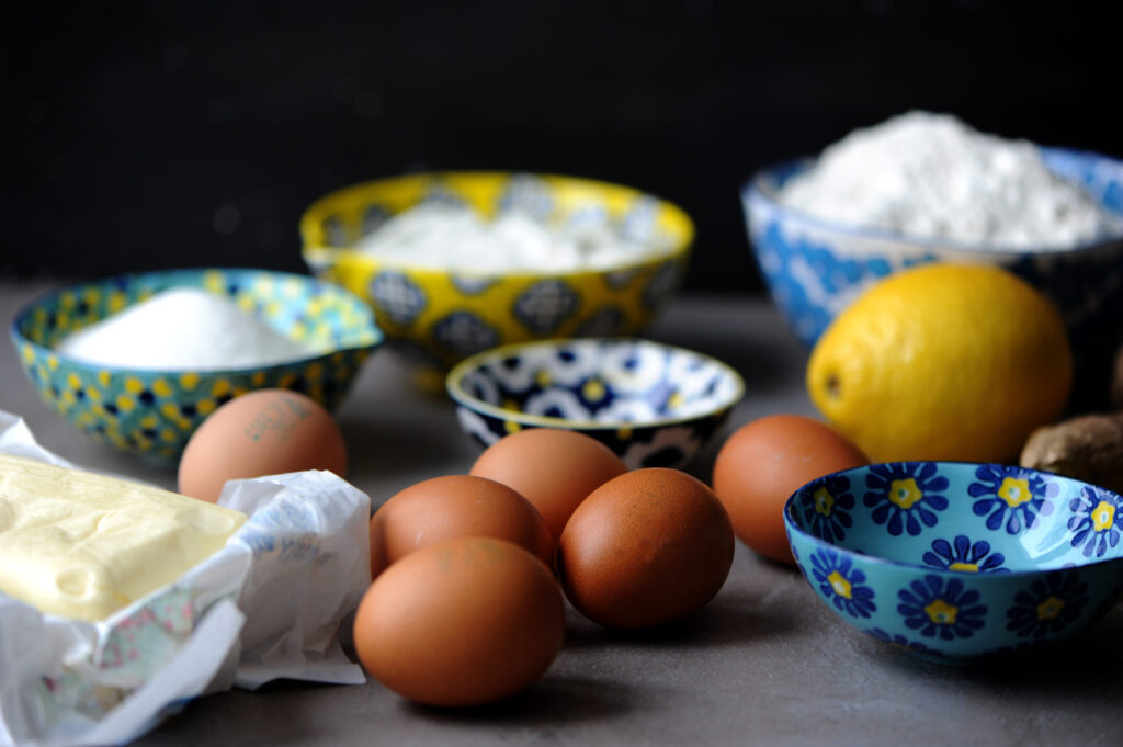 Close-up of raw baking ingredients for a lemon bundt cake, showing brown eggs, butter, a fresh lemon, and flour in colorful patterned bowls.