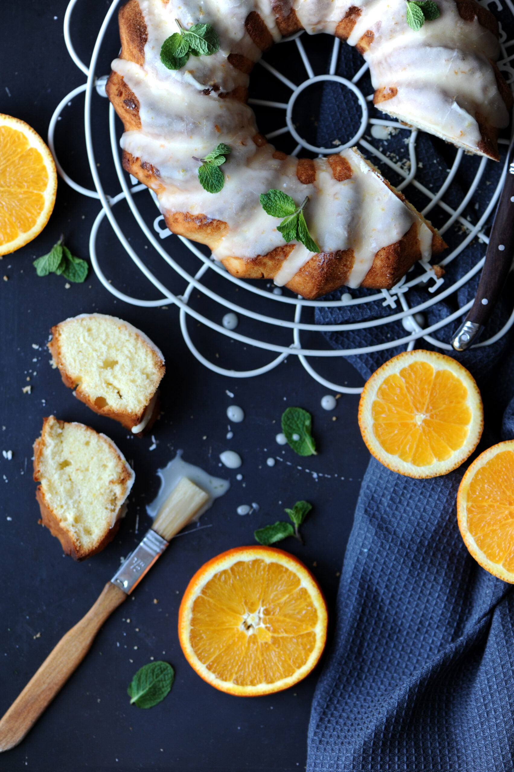 Top-down view of sliced lemon ginger bundt cake with white glaze, fresh orange slices, a pastry brush, and a blue napkin on a dark background.
