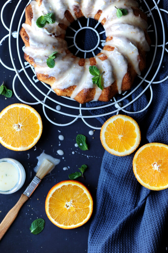 Overhead view of a glazed lemon ginger bundt cake on a wire rack, surrounded by fresh orange slices, a bowl of glaze, a pastry brush, and a blue napkin on a dark surface.