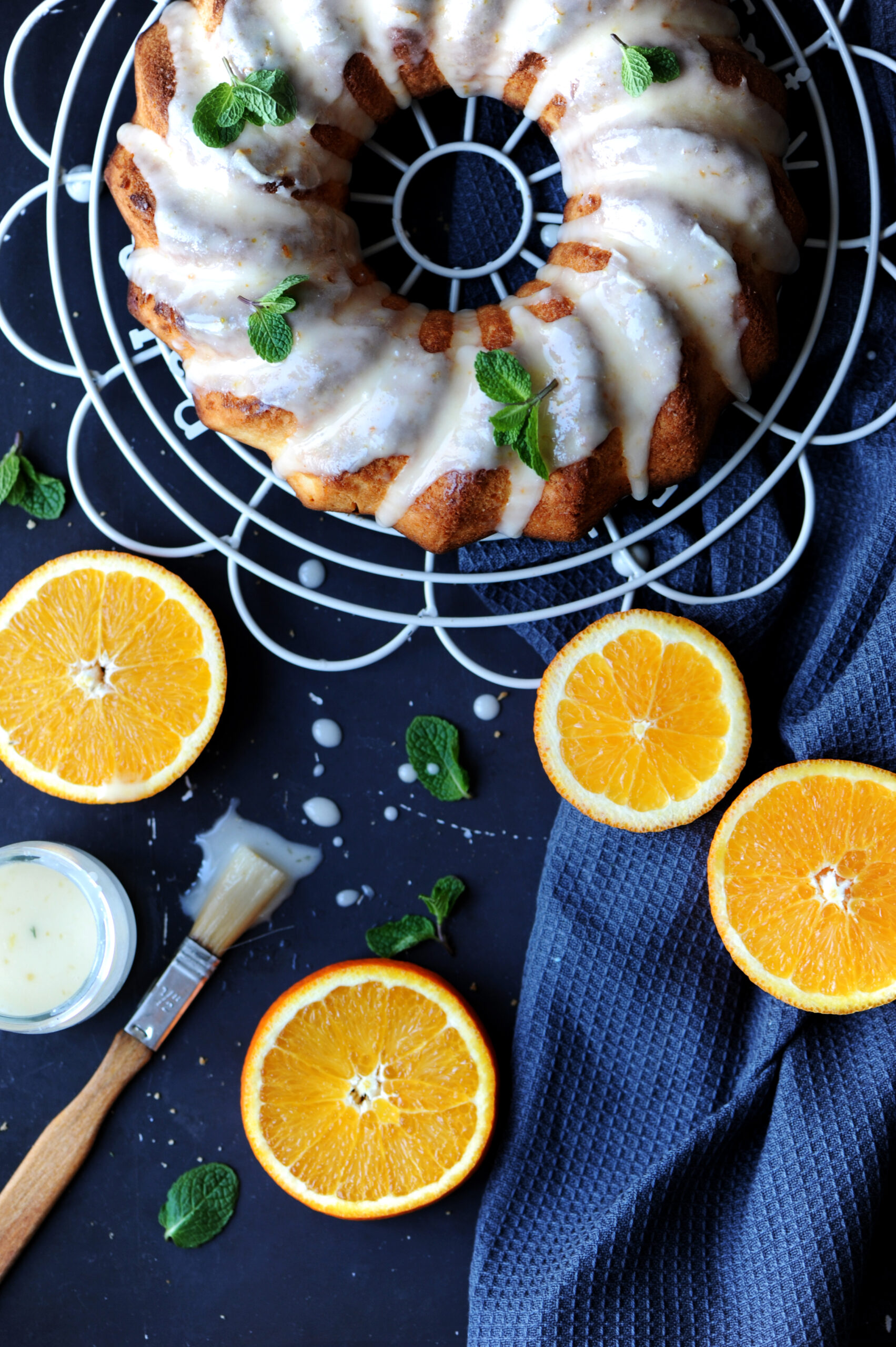 Overhead view of a glazed lemon ginger bundt cake on a wire rack, surrounded by fresh orange slices, a bowl of glaze, a pastry brush, and a blue napkin on a dark surface.