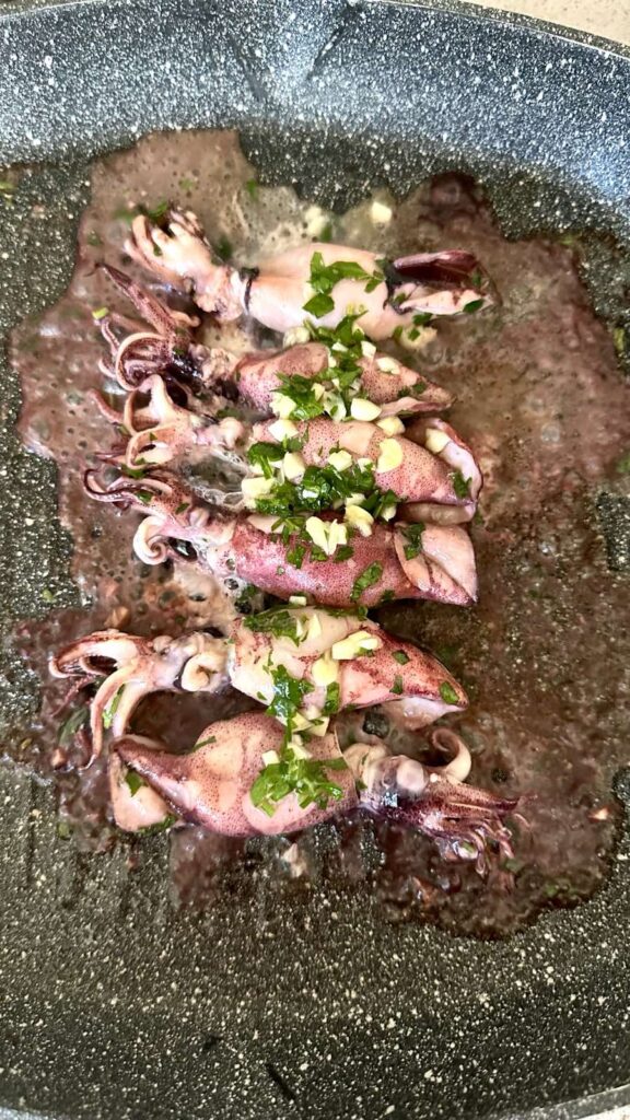Baby squid cooking in a speckled hot pan topped with minced garlic and fresh green parsley