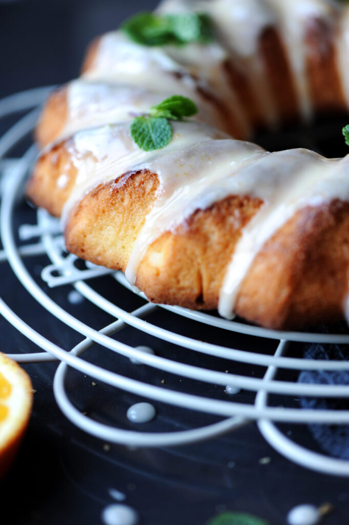 A whole lemon ginger bundt cake drizzled with white orange glaze and garnished with fresh mint on a wire cooling rack.
