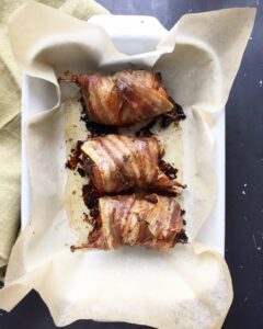 Oven-baked bacon-wrapped quail stuffed with prunes resting in a white baking dish on roasted parchment paper.