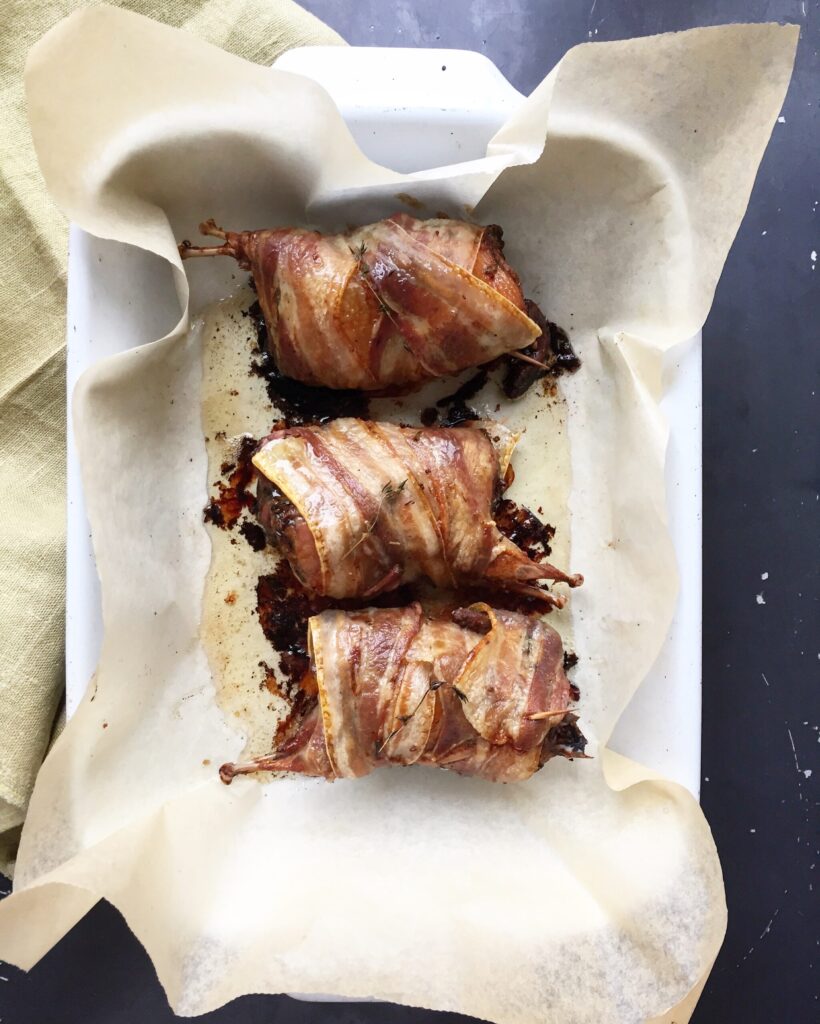 Oven-baked bacon-wrapped quail stuffed with prunes resting in a white baking dish on roasted parchment paper.