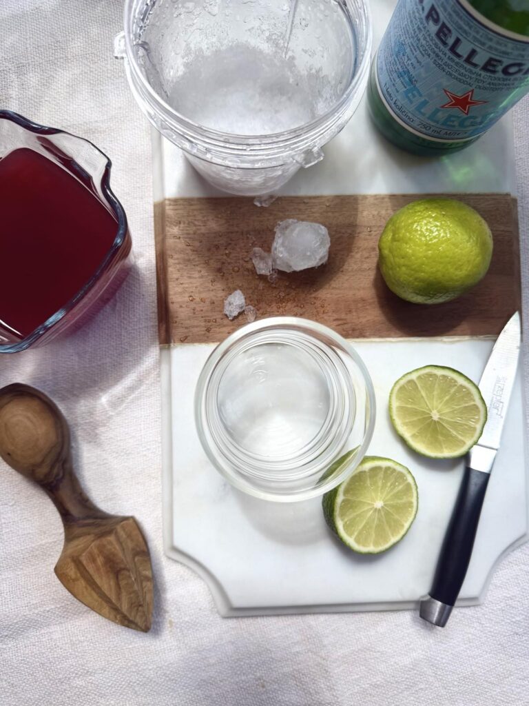 Ingredients for pomegranate limeade, including pomegranate juice, sliced limes, ice cubes, and a bottle of San Pellegrino sparkling water
