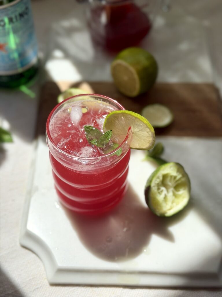 Glass of pomegranate limeade garnished with a lime wheel and fresh mint leaves, surrounded by halved limes