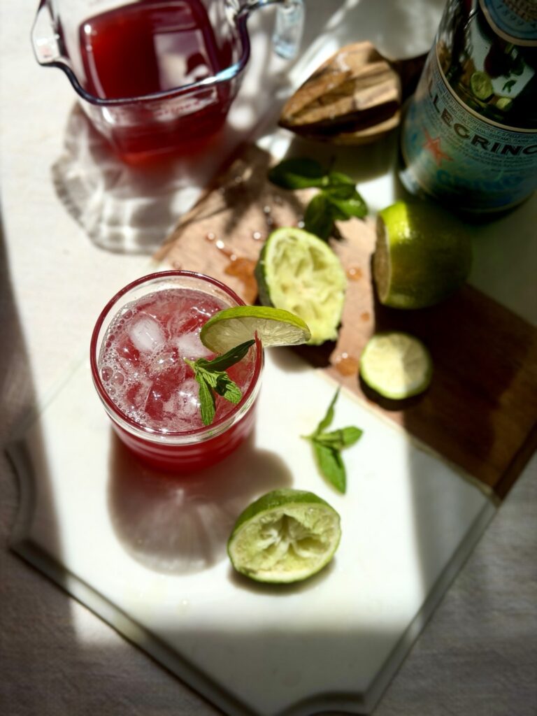 Glass of pomegranate limeade garnished with mint and lime next to a bottle of San Pellegrino sparkling water