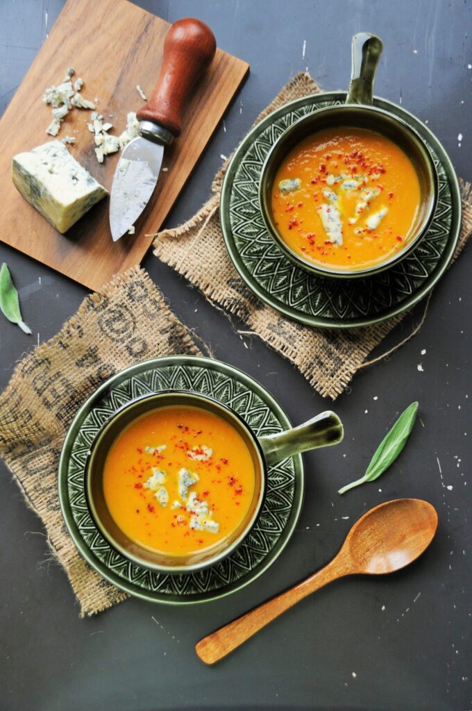 Two green bowls of creamy pumpkin soup topped with crumbled blue cheese and fresh sage, served on a dark rustic background with a wooden spoon.