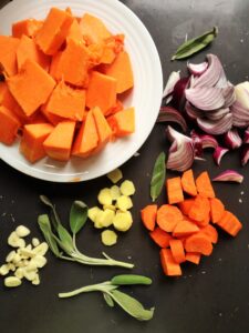 A top-down view of raw ingredients for pumpkin soup on a dark surface, featuring chopped orange pumpkin in a white bowl, sliced red onions, diced carrots, sliced ginger, garlic cloves, and fresh sage leaves.