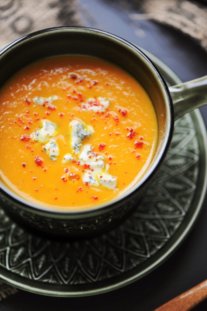 A close-up shot of a dark ceramic bowl filled with creamy orange pumpkin soup, garnished with crumbled blue cheese, a drizzle of cream, and red chili flakes, sitting on a matching saucer.