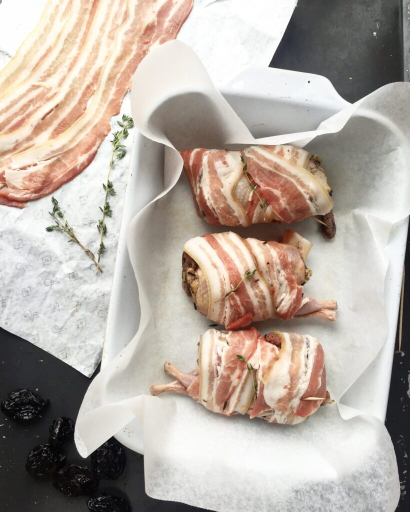 Raw bacon-wrapped quail stuffed with prunes placed in a parchment-lined white baking dish before baking.