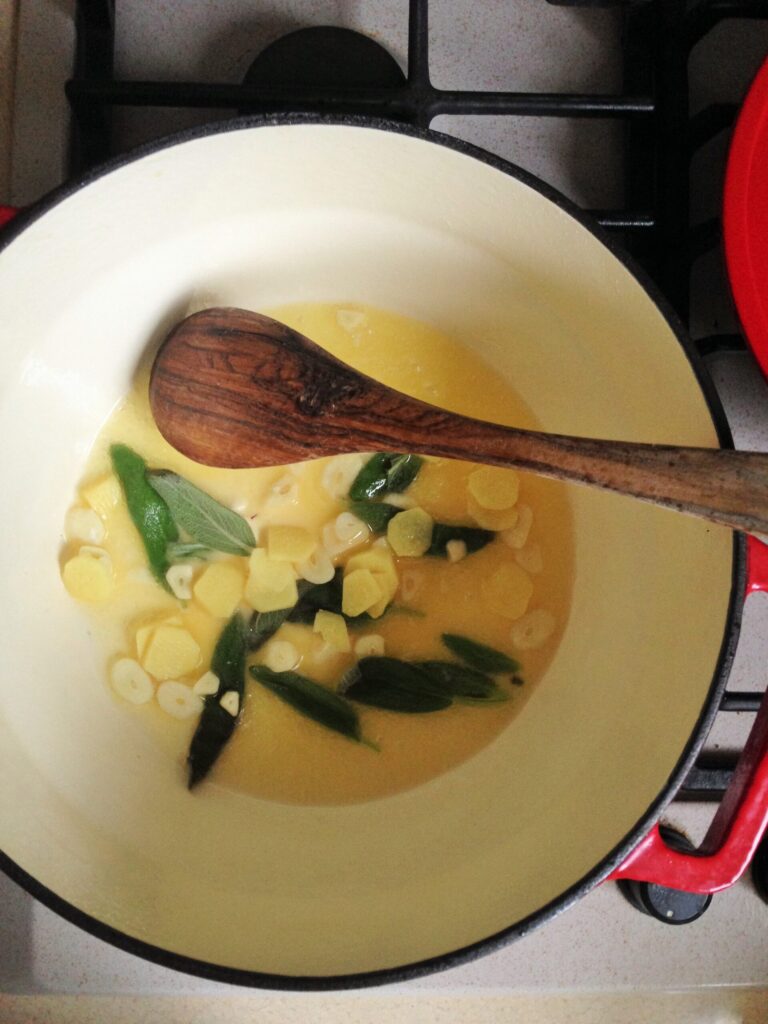 A top-down view of sliced garlic, chopped ginger, and fresh sage leaves being sautéed in oil inside a pot with a wooden spoon.