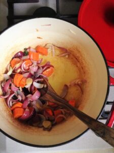 A top-down view of chopped red onions and carrots being sautéed in oil inside a light-colored pot with a wooden spoon.