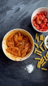 Bowl of seasoned raw chicken pieces next to chopped tomatoes and dry fusilli pasta on a dark background