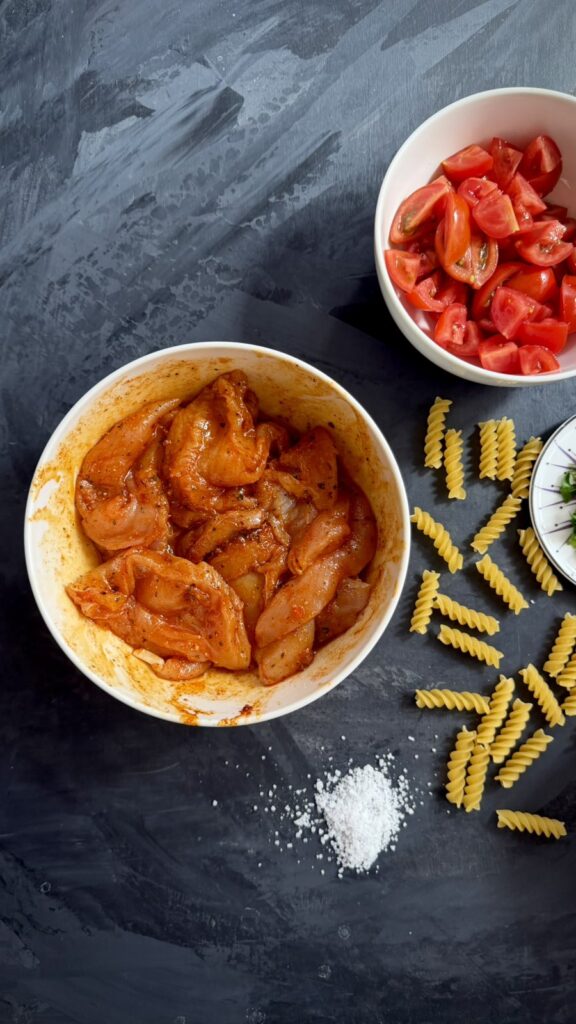 Bowl of seasoned raw chicken pieces next to chopped tomatoes and dry fusilli pasta on a dark background