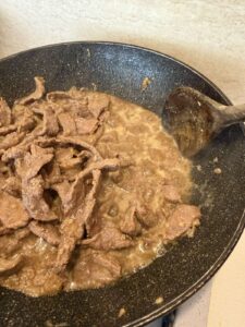 Beef strips simmering in soy sauce marinade in a skillet, being stirred with a wooden spoon