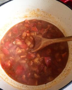 Simmering tomatoes, red bell peppers, and onions in a pot with a wooden spoon, creating the base for a creamy tomato and red pepper soup.