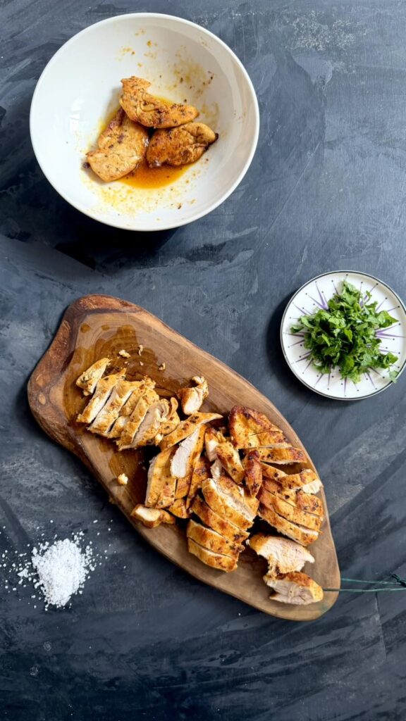 Cooked chicken breast sliced on a wooden board next to a bowl of fresh herbs
