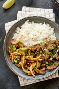Plated soy sauce beef stir-fry served with steamed rice and a spicy cucumber salad, garnished with green onions