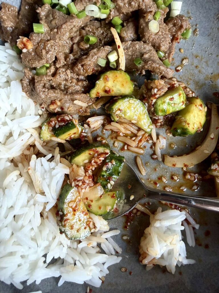 Overhead close-up of soy sauce beef, cucumber salad, and rice on a plate, garnished with green onions and sesame seeds
