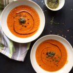 Two white bowls of creamy tomato and red pepper soup garnished with a dollop of pesto, fresh thyme, and red pepper flakes on a dark surface.