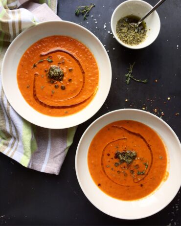 Two white bowls of creamy tomato and red pepper soup garnished with a dollop of pesto, fresh thyme, and red pepper flakes on a dark surface.
