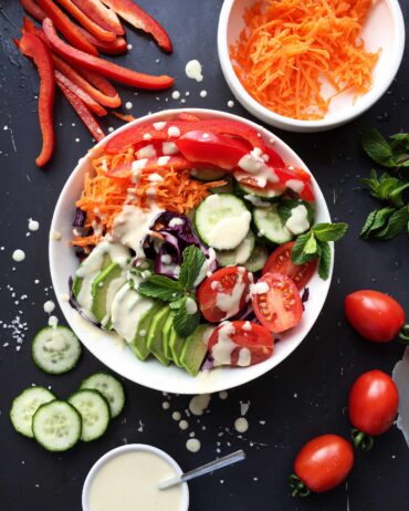 Rainbow vegetable salad bowl with sliced avocado, tomatoes, cucumbers, carrots, and creamy hummus dressing on a dark background.