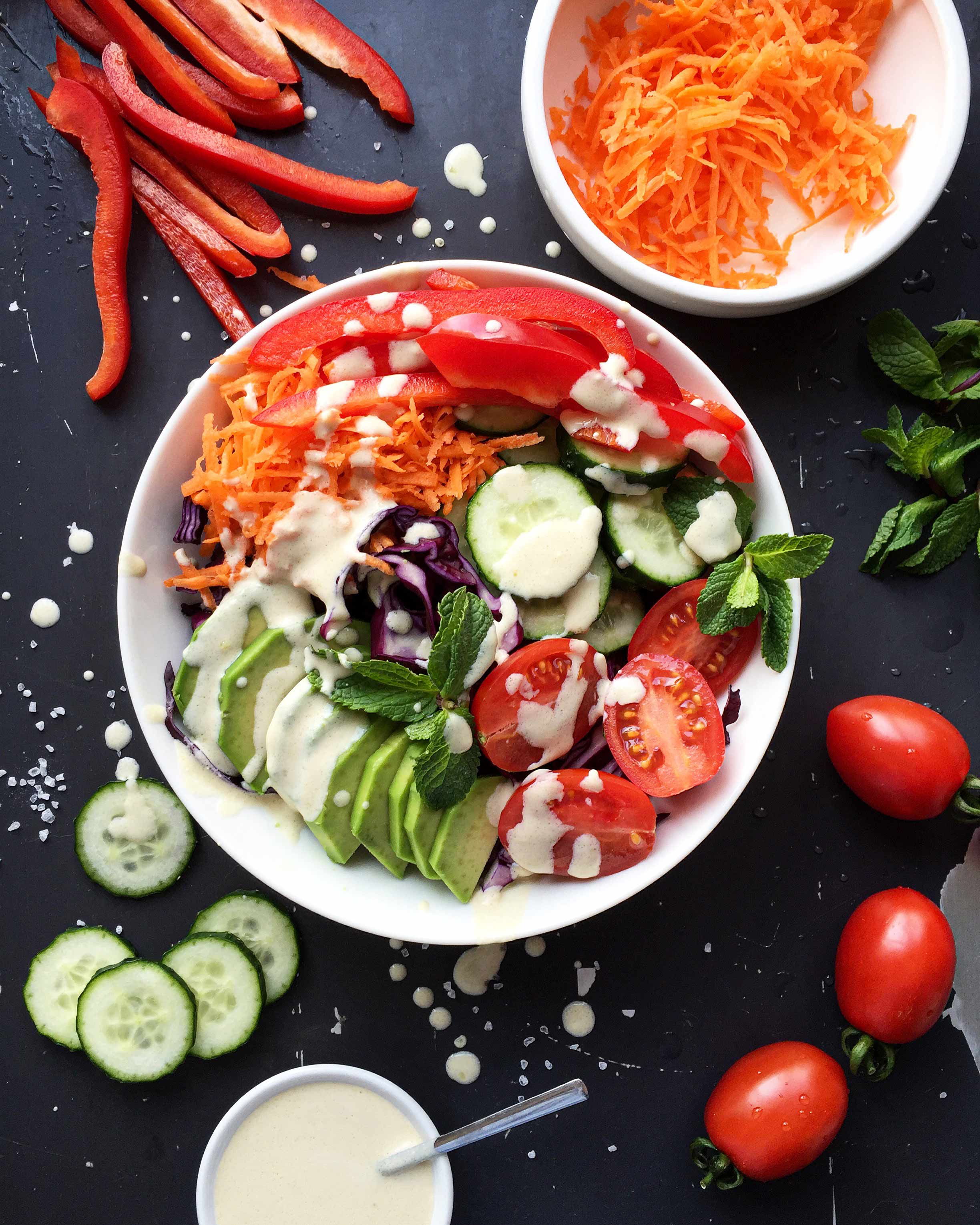 Rainbow vegetable salad bowl with sliced avocado, tomatoes, cucumbers, carrots, and creamy hummus dressing on a dark background.