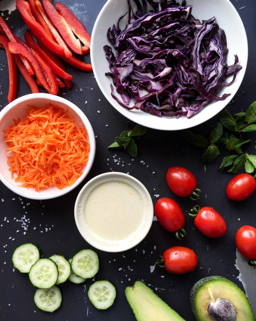 Fresh ingredients for a rainbow vegetable salad bowl including sliced red bell peppers, shredded carrots, tomatoes, and cucumbers.