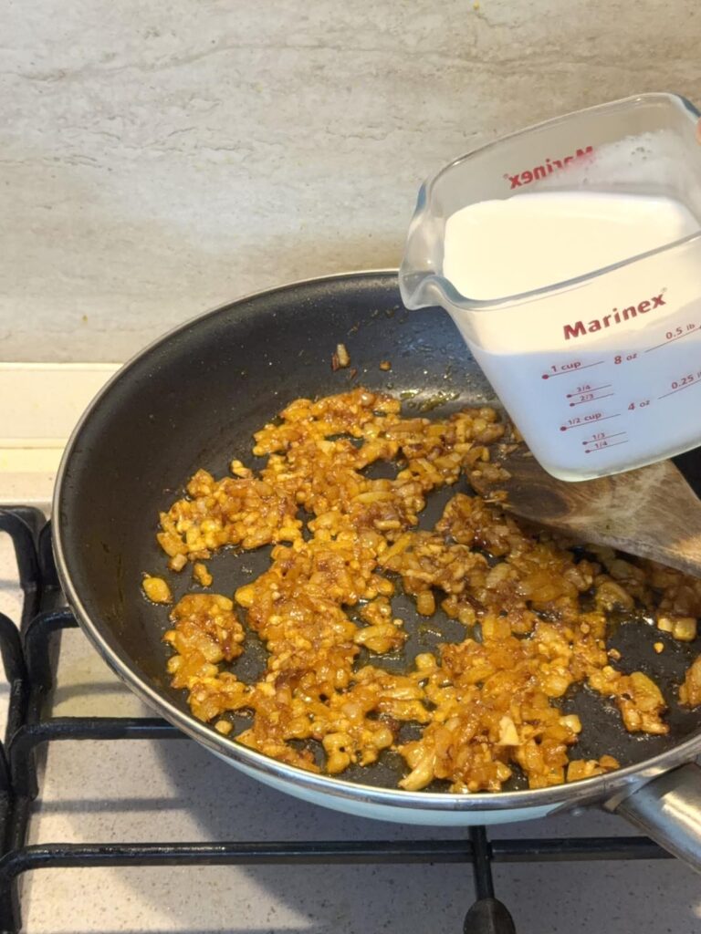 Pouring coconut milk into a skillet with sautéed onions and Tom Yum paste.