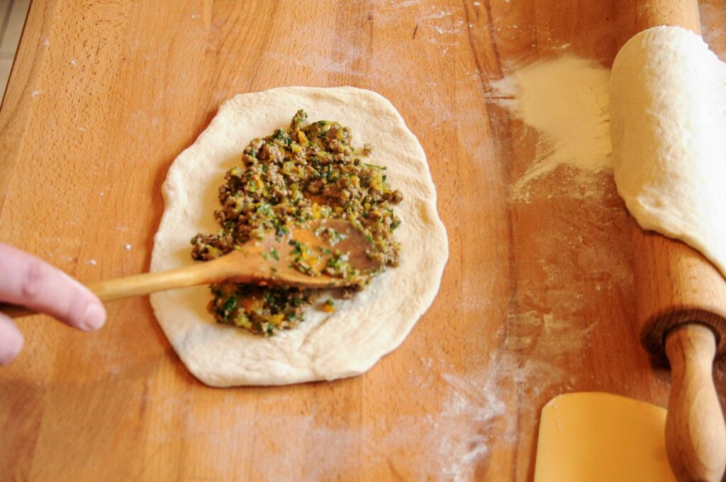 A hand using a wooden spoon to add a ground beef and vegetable filling to a circle of raw Turkish pide dough on a flour-dusted wooden board