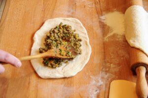 A hand using a wooden spoon to add a ground beef and vegetable filling to a circle of raw Turkish pide dough on a flour-dusted wooden board