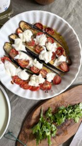 Top-down view of two white plates serving roasted eggplant halves topped with mozzarella, tomatoes, and fresh herbs, arranged on a grey linen tablecloth alongside a glass of white wine.