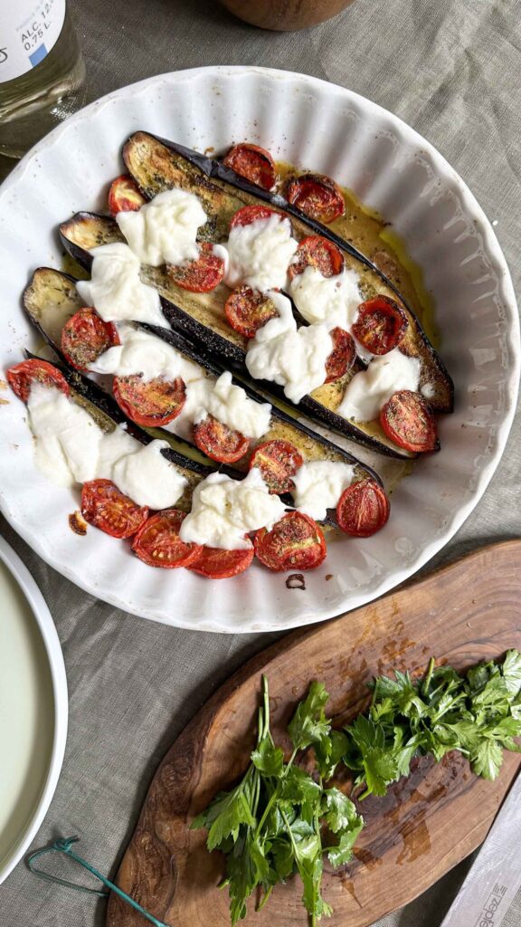 Top-down view of two white plates serving roasted eggplant halves topped with mozzarella, tomatoes, and fresh herbs, arranged on a grey linen tablecloth alongside a glass of white wine.