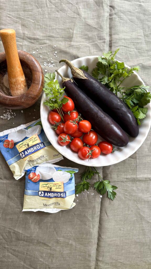 Ingredients for baked eggplant with mozzarella and tomatoes: raw eggplant halves, fresh cherry tomatoes, mozzarella cheese, and herbs on a linen cloth.