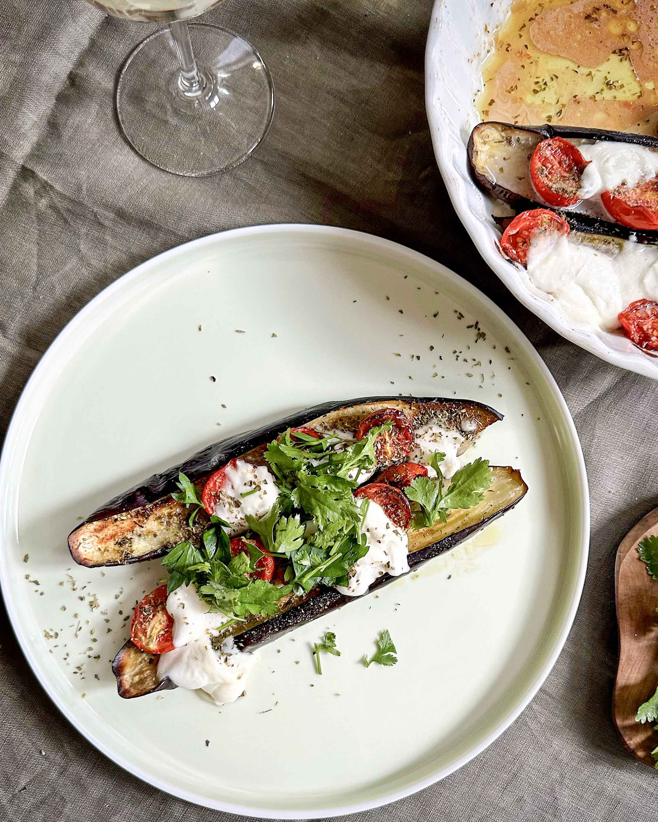 Plated baked eggplant halves topped with melted mozzarella, roasted cherry tomatoes, and fresh herbs on a white ceramic plate.