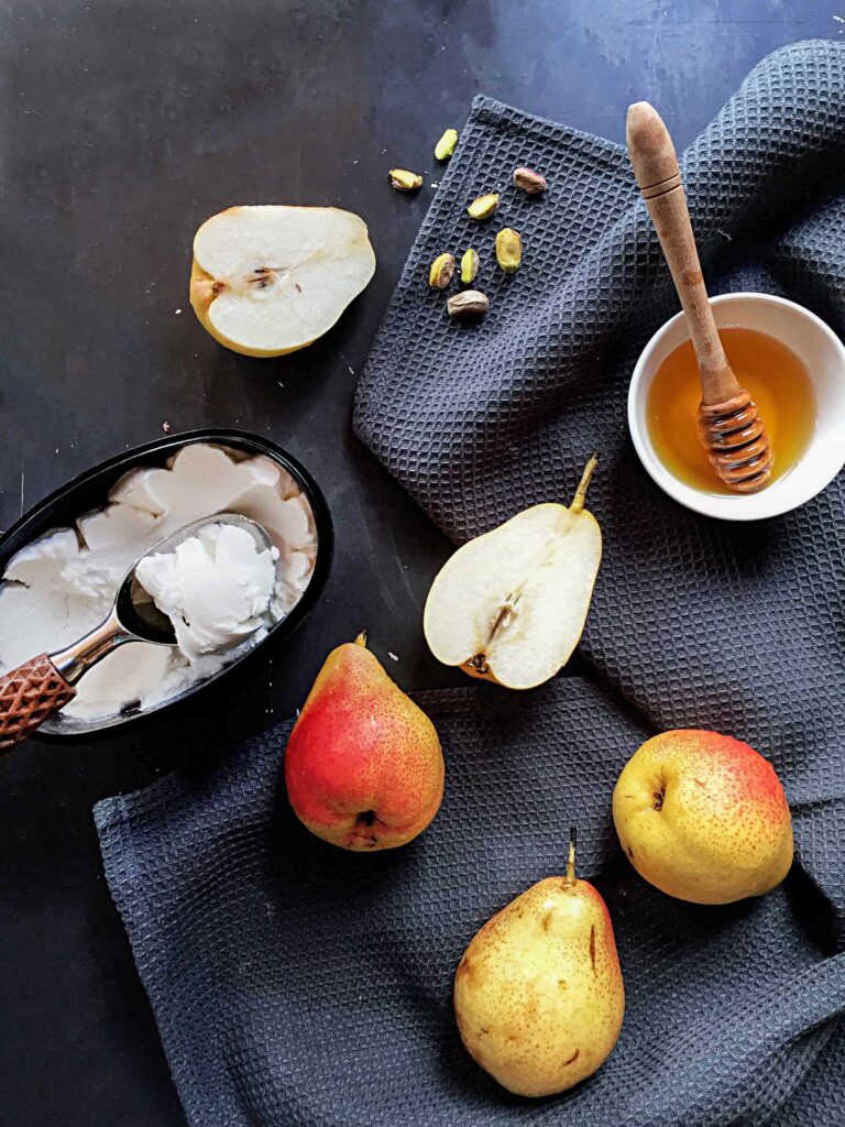 A flat lay of ingredients for a baked pear dessert featuring whole and halved pears, a bowl of honey with a wooden dipper, vanilla ice cream with a spoon, and scattered pistachios on a dark linen cloth.