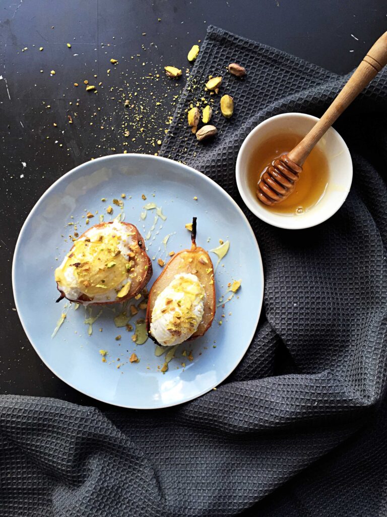 Top-down view of two baked pear halves topped with vanilla ice cream and crushed pistachios on a light blue plate, set on a dark gray linen napkin with a bowl of honey and a wooden dipper nearby.