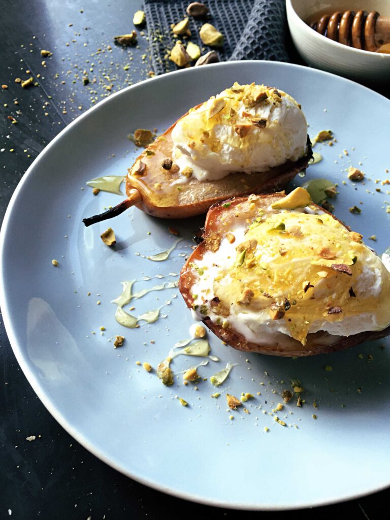 Two warm baked pear halves topped with melting vanilla ice cream, crushed pistachios, and honey, served on a light blue plate with a honey dipper in the background.