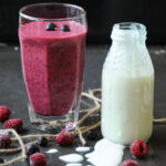 A tall glass of black currant and raspberry smoothie next to a small glass bottle of yogurt on a dark surface with scattered fresh berries