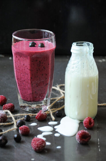 A tall glass of black currant and raspberry smoothie next to a small glass bottle of yogurt on a dark surface with scattered fresh berries