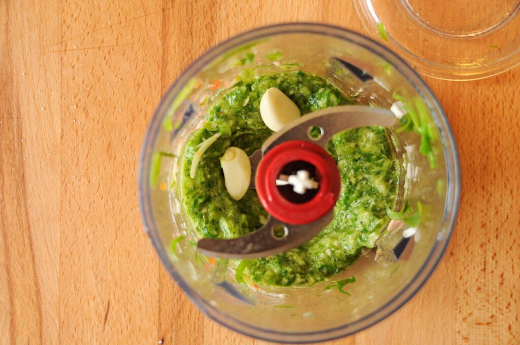 Top-down view of a food processor bowl containing a bright green blended herb mixture and whole garlic cloves on a wooden board