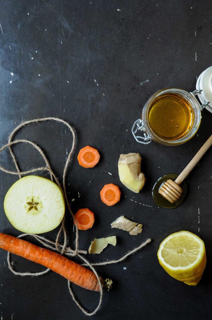 A top-down view of fresh juice ingredients on a dark surface, featuring a halved green apple, fresh carrots wrapped in rustic twine, sliced carrots, ginger pieces, a lemon half, and a jar of honey with a wooden dipper.
