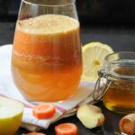 A glass of fresh, vibrant orange carrot, apple, and ginger juice resting on a white napkin, surrounded by raw ingredients including green apple wedges, carrot slices, ginger root, a lemon half, and a small jar of honey on a dark rustic surface.