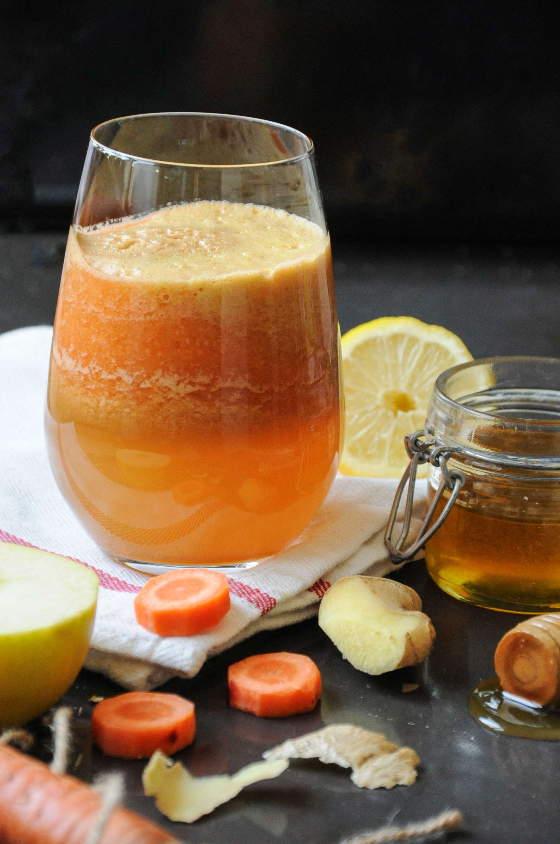 A glass of fresh, vibrant orange carrot, apple, and ginger juice resting on a white napkin, surrounded by raw ingredients including green apple wedges, carrot slices, ginger root, a lemon half, and a small jar of honey on a dark rustic surface.