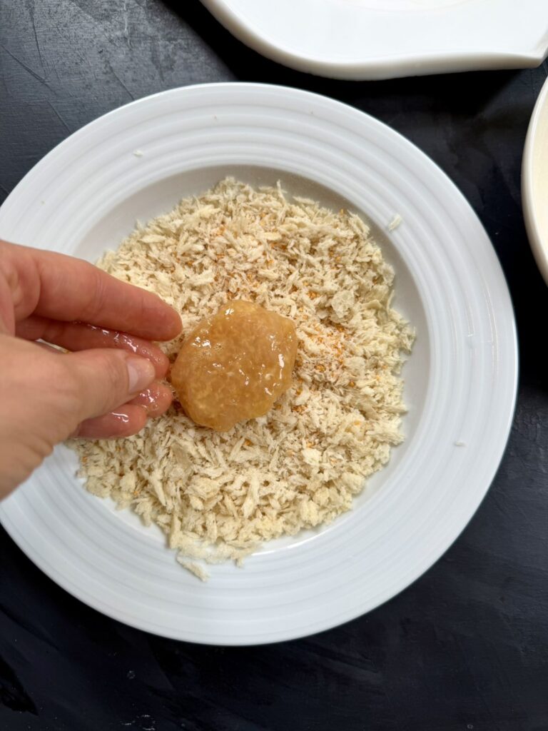 A hand pressing a nugget into a bowl of breadcrumbs.