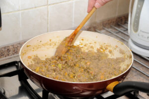 A hand using a wooden spoon to stir a steaming mixture of ground beef and diced vegetables in a red skillet on a gas stovetop